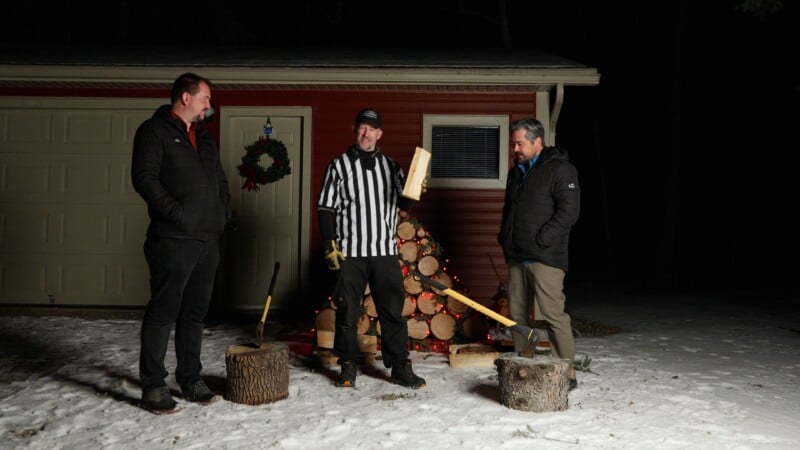 Three men stand outside on snow in front of a red garage at night. One wears a referee shirt and holds a log, with axes and chopped wood nearby. A stack of firewood decorated with lights is in the background.