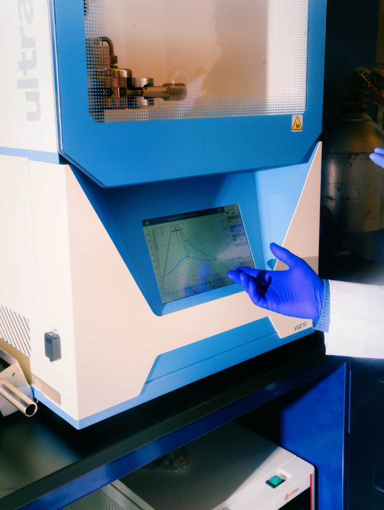 A lab technician’s gloved hand gestures to a piece of equipment — a large white box with a window showing metal machinery in the top half and a display screen in the bottom half.