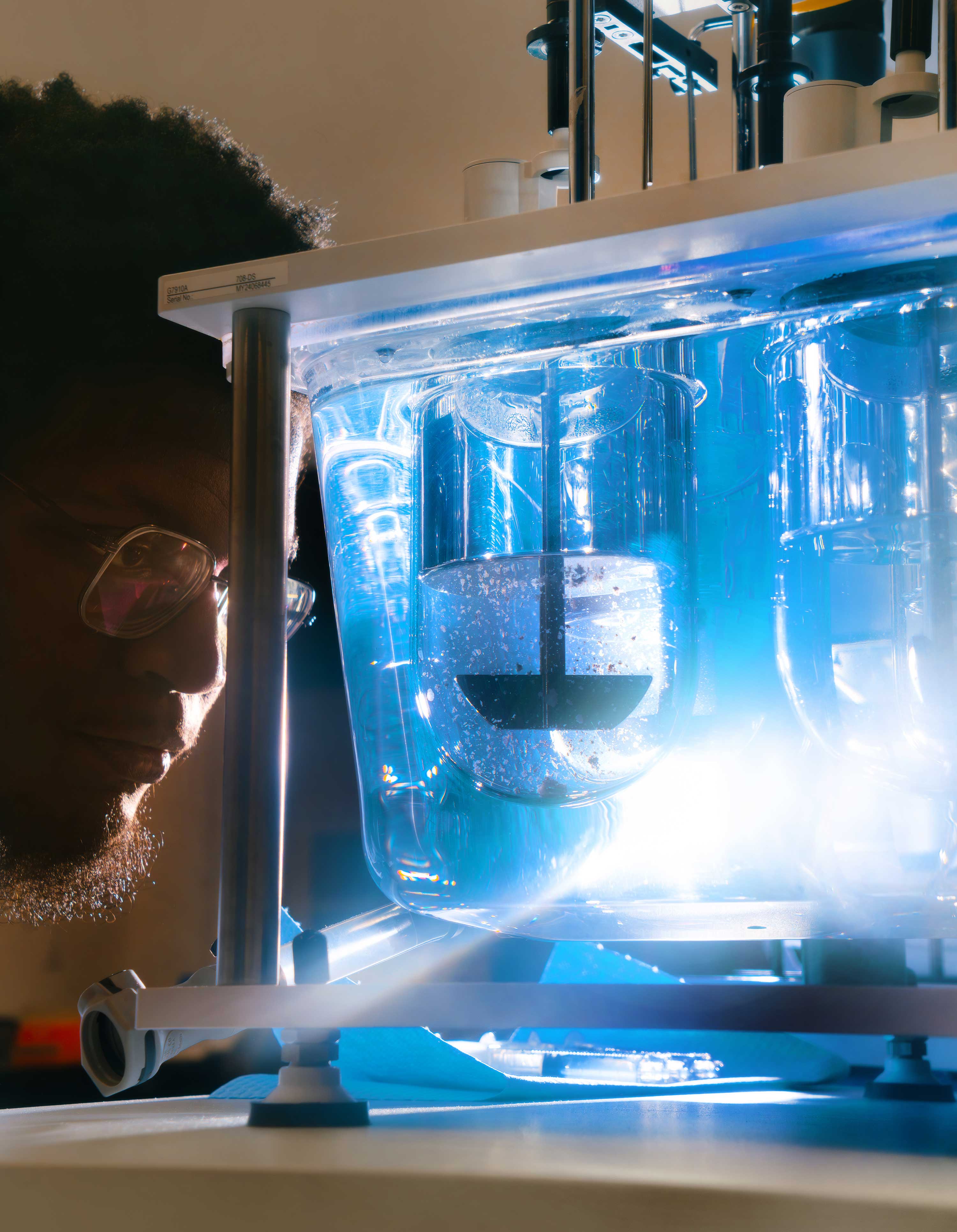 A lab technician leans in close to observe a drug tablet dissolving in a glass container of liquid.