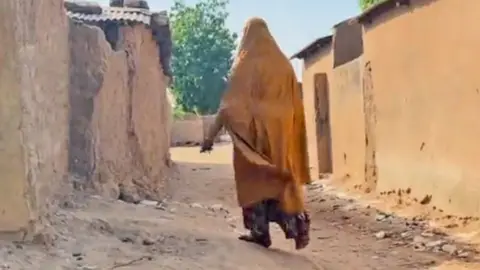 Gift Ufuoma/BBC A woman wearing a mustard-coloured robe, seen from behind, walking through a  street in Nukuru with mud-walled buildings on either side.
