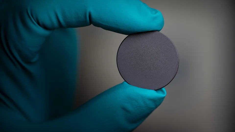 A close-up of a gloved hand holding a small, circular grey disc - which is a sphere of material used in semiconductors. The glove is blue and the disc is being held between two fingers. 