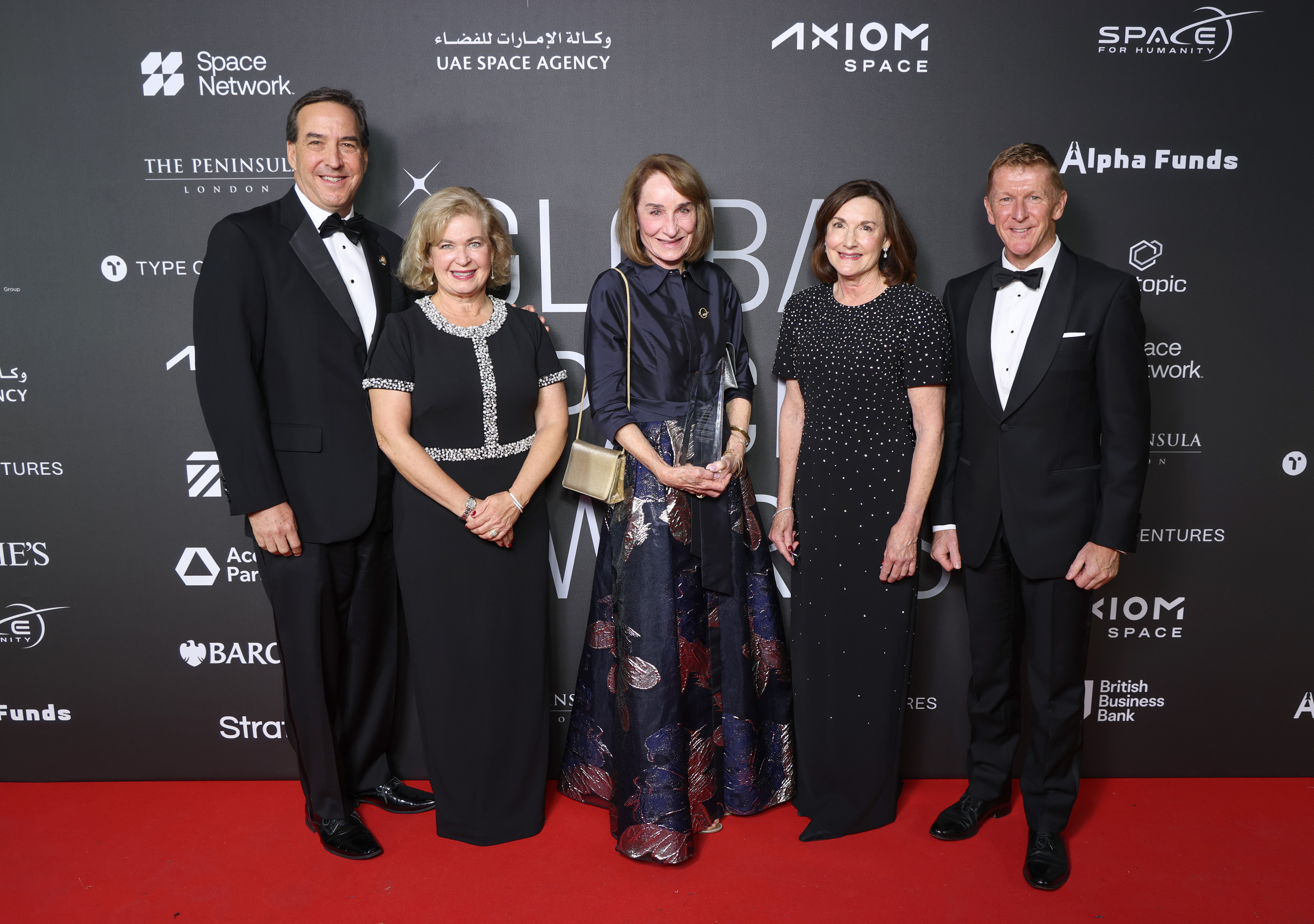 A group of five people in formal tuxedos and dresses stand on a red carpet.