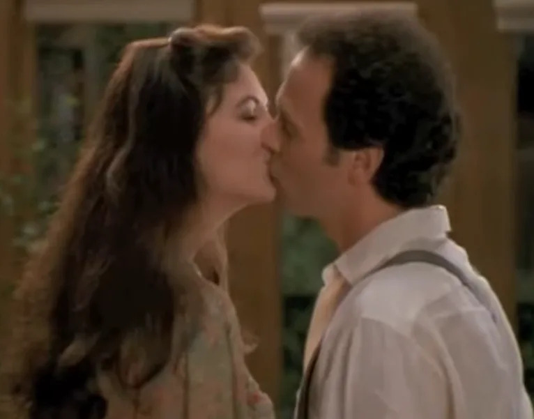 Two people are kissing in an indoor setting, with one wearing a shirt and suspenders