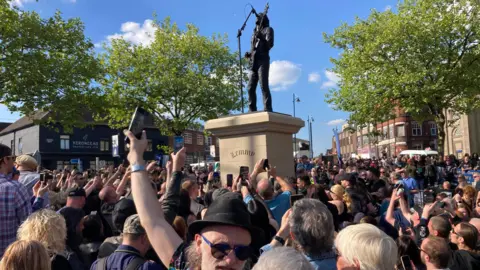 A crowd of people gathered in a town square around the statue of Lemmy Kilmister. Many are holding up phones.