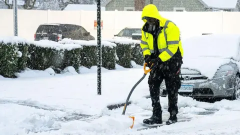 Getty Images A man in a fluorescent jacket and black trousers and boots shovels snow by the side of a road