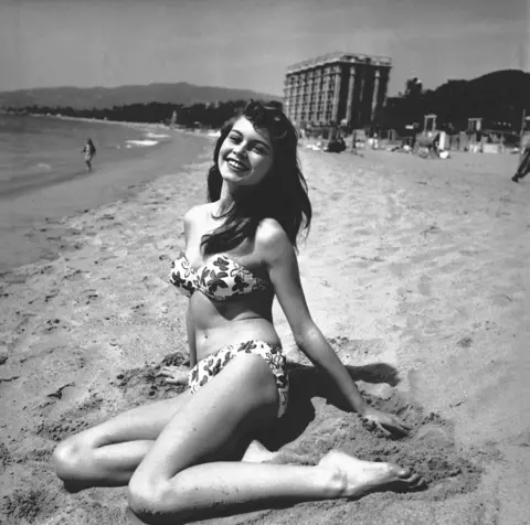 Lido/Shutterstock Brigitte Bardot pictured in black-and-white, with long dark hair, wearing a floral bikini as she sits on the sand smiling for photographers at the Cannes Film Festival in 1953.