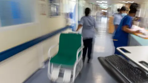 PA Media A busy ward in a hospital. One member of staff in uniform is pulling a green chair. The background is blurred. 