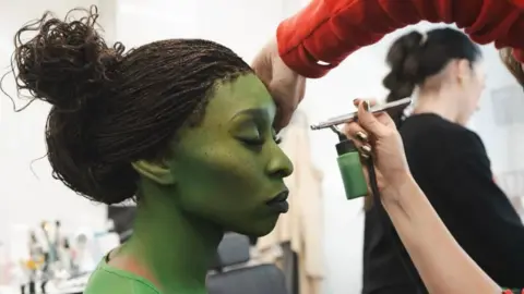 Andres Di Rienzo Cynthia Erivo sat in a make-up chair in a make-up trailer on the set of Wicked. She has her eyes closed and is being painted green by someone who is using an air brush machine. 