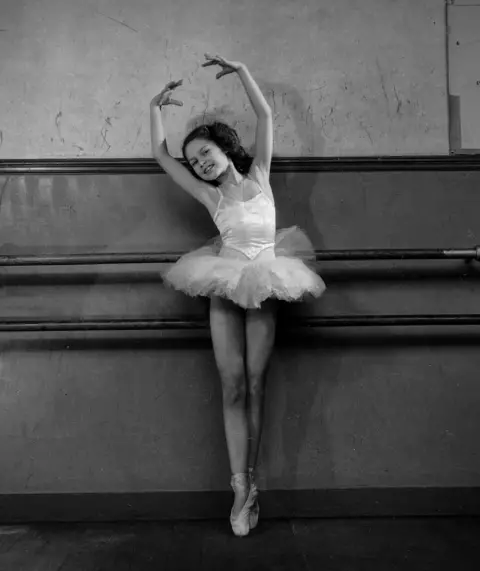 Roger Viollet via Getty Images Brigitte Bardot, pictured in a black-and-white photo from about 1946, in a ballerina's tutu, standing en pointe with her arms raised and leaning to her left.