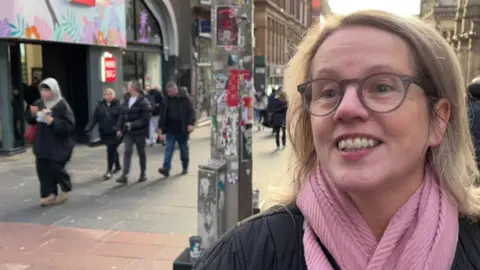 A shopper with short blonde hair, glasses, a pink scarf, smiling and wearing a black jacket.