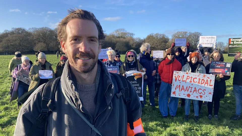 Laurence Bradley stood in front of a group of protesters holding up signs. He has short brown hair and facial hair and is wearing a black coat. The sky is blue.