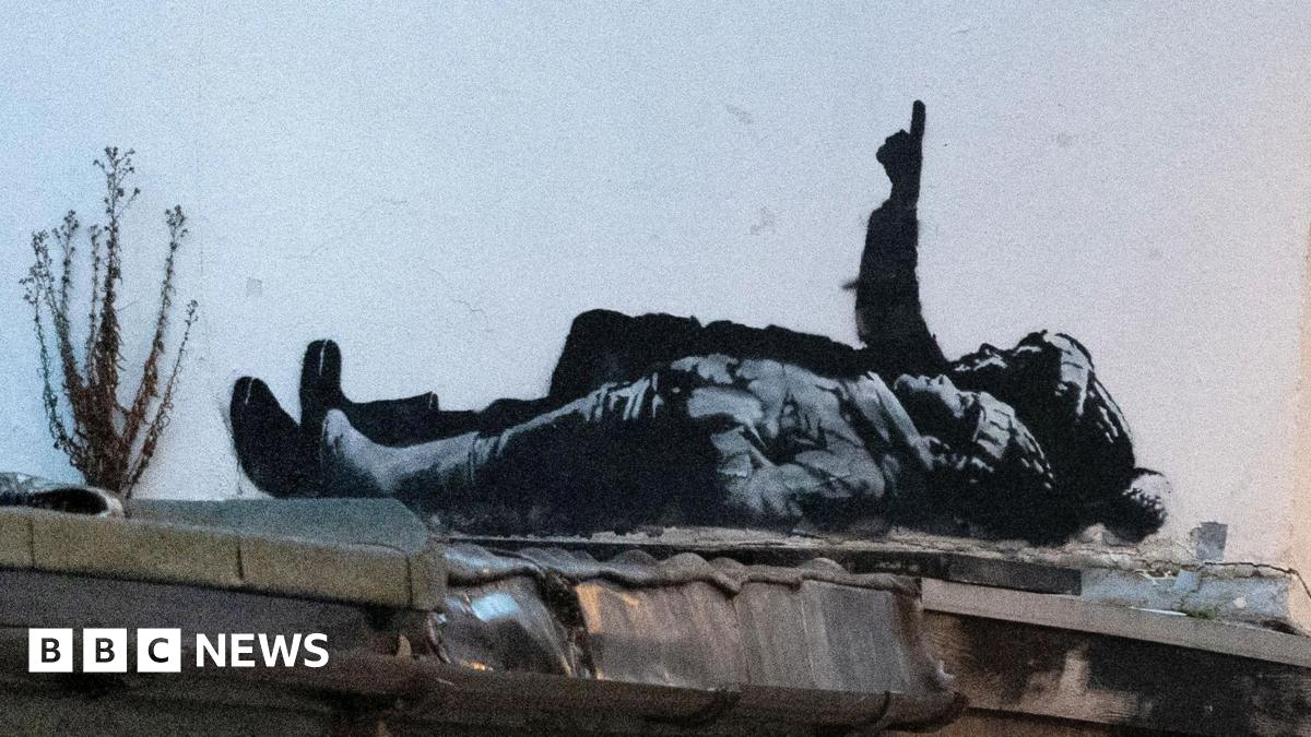 The black and white street art depicts two children lying on their backs and pointing upwards. The child nearest the camera is wearing a wooly hat with hands tucked into their jacket pockets. Behind them a larger person is pointing upwards in a bobbly hat (possibly a Santa type hat) also wearing boots.