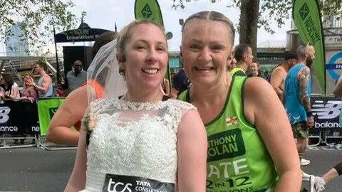 Chloe Morgan Mrs Coleman-Day and Mrs Walford stood side-by-side after completing the marathon. Mrs Coleman-Day has a white wedding dress on and Mrs Walford is wearing black and green running gear. 
