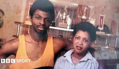 A young James Cook, wearing a yellow boxing vest, with his arm around his wife Carmen. There are various sport trophies behind them.