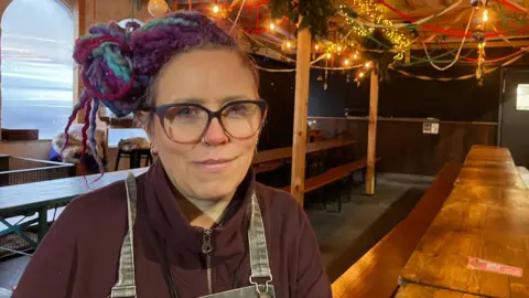 Sarah Shearman sits on a bench at a long table that is lit by fairy lights and has Christmas decorations. Sarah has blue, purple and red coloured hair tied up to the side, a purple fleece and denim dungarees.