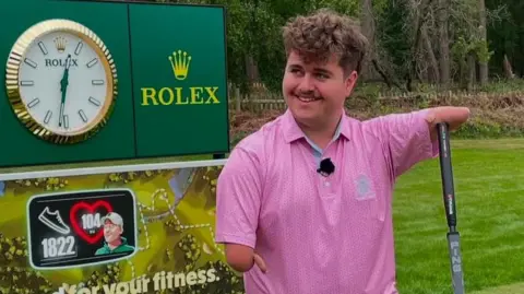 Adam Dalton has under-developed arms. He is holding onto a golf club with his arm and wearing a pink golf shirt.