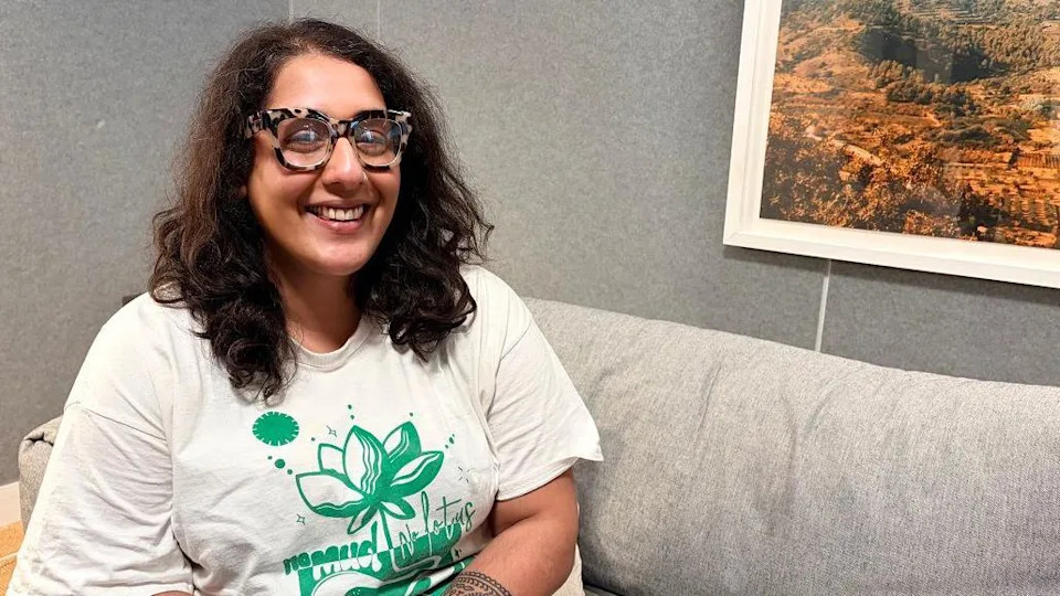 Corrina is sitting on a grey sofa and there is a picture on the wall behind her. She has long, curly black hair and glasses and is wearing a green and white T-shirt.