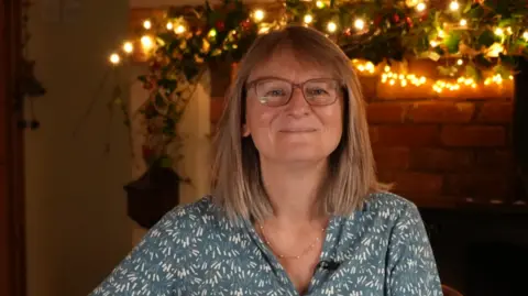 Shaun Whitmore/BBC A woman with mid length dark blonde hair smiles at the camera in a head and shoulders image. She is wearing brown-framed spectacles and a teal and cream-patterned blouse. 
