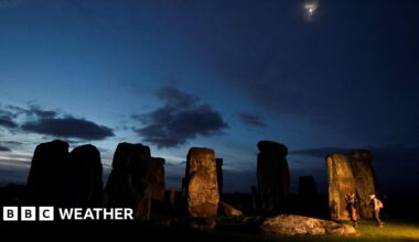 Stonehenge at dawn, with a mainly dark sky with glimmers of brightness and the moon overhead and two people next to the standing stones.
