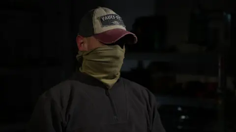Kevin McGregor/BBC Ruslan, Special Operations Forces  with a hat and snood hiding his face and identity in a dark room wearing a black fleece