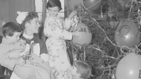Getty Images A black-and-white image of a nurse holding up a child in a dressing gown to place a fairy on top of a Christmas tree. Another child looks on at her feet and a third in a dressing gown is standing on something as she hangs balloons on the tree.