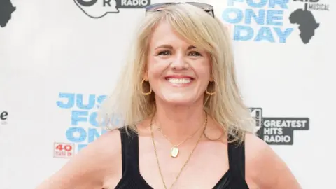 PA Media Sally Lindsay smiles at the camera. She has long blonde hair and wears gold hoop earrings two gold necklaces and a black sleeveless dress. 