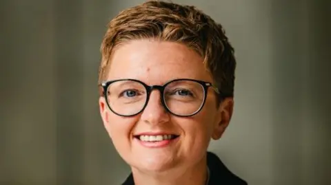 Manchester City Council Councillor Bev Craig smiles at the camera. She has short, light brown hair and wears round, black-rimmed glasses
