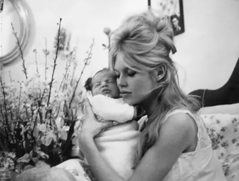 AFP via Getty Images Brigitte Bardot, wearing a nightdress with her blonde hair in a beehive hairdo, holding her son close to her face.