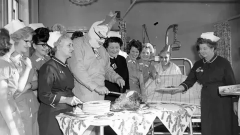 Getty Images A black-and-white image of a man carving a turkey, surrounded by women in nurse's uniforms. The man is wearing overalls, bandages over his face and a star on his head as he carves the turkey on a table covered in a decorative festive cloth. A nurse holds out a plate for the turkey to be served. A male patient can be seen sitting up in a bed in the background, wearing a hat. All are smiling.