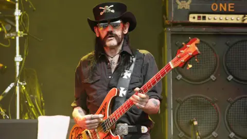 PA Media Lemmy from Motörhead wearing a black hat, black shirt and holding an orange coloured bass guitar, performs on stage. He is standing in front of a large amplifier.