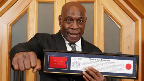 PA Media Frank Bruno holding his framed certificate showing his freedom of the City of London. His right hand is holding his fist in a boxing punch toward camera. 