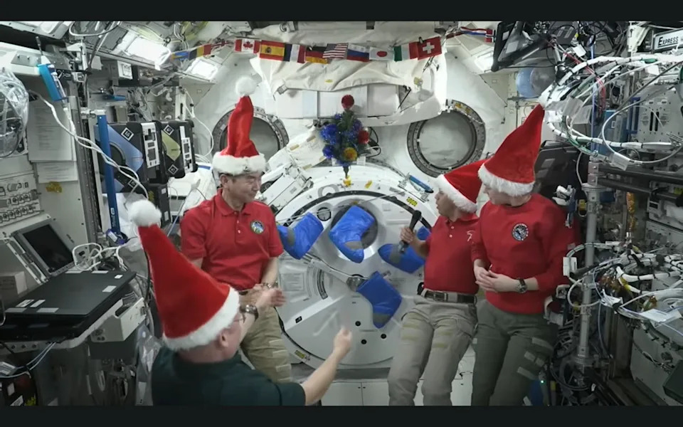 Four astronauts in Santa hats inside a space station decorated with space stockings and a Christmas tree.