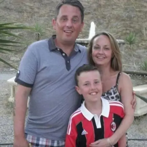 Mark Williams A man in a blue t-shirt and checked shorts, with his arm round a blonde woman in a sundress. In front of them is a young brown-haired boy wearing a black and red sports shirt. They are all looking at the camera and smiling.