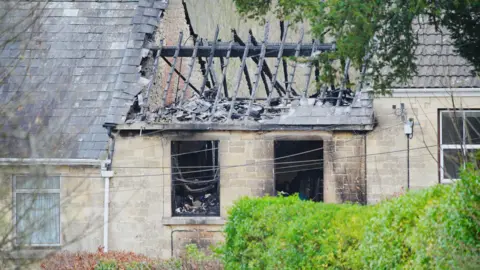 PA The charred shell of the destroyed home following a fire. The roof has collapsed and only the burnt wooden beams are visible. The glass windows have been smashed and are completely blackened from smoke. The surrounding homes on the terrace are untouched.