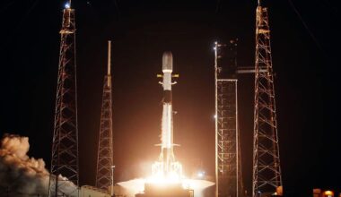 a white and black rocket lifts off into the darkness of night, its bright orange-tinted plume lighting up its launch pad.