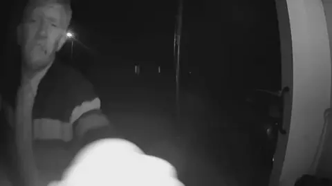 Supplied Night-vision doorbell footage showing a middle-aged blonde or white-haired man, with a cigarette in his mouth and wearing a tracksuit jacket, reaching towards camera.
