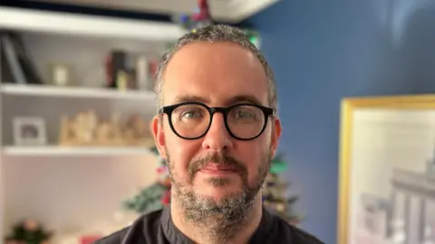 BBC Keith Lawrence is a man with black and white hair and beard. He is wearing black glasses and a black grandad collar shirt. He is standing in his sitting room which is out of focus in the background. The walls are blue, there is a white cabinet, a white painting on the wall and a Christmas tree directly behind him.