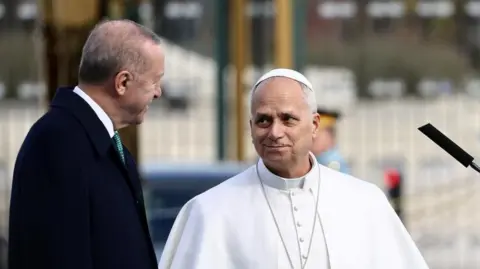 Getty Images A man in white clerical garb talks to a man in a black business coat. 