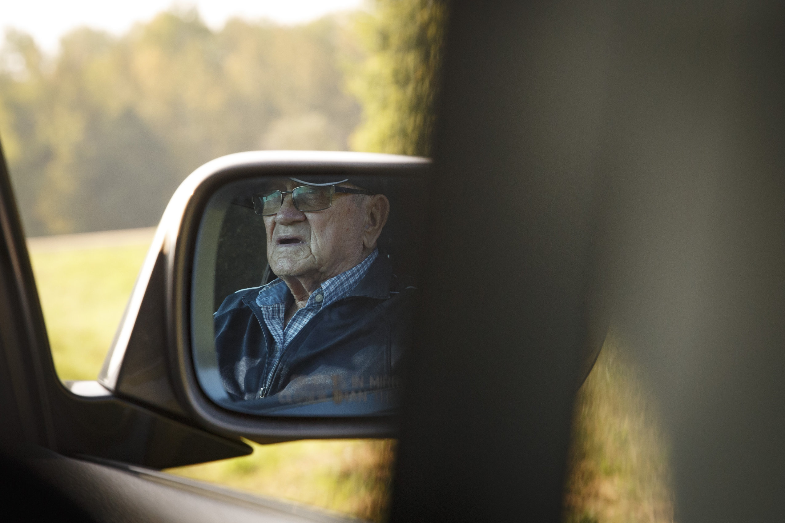 An older man with glasses is reflected in a car mirror.