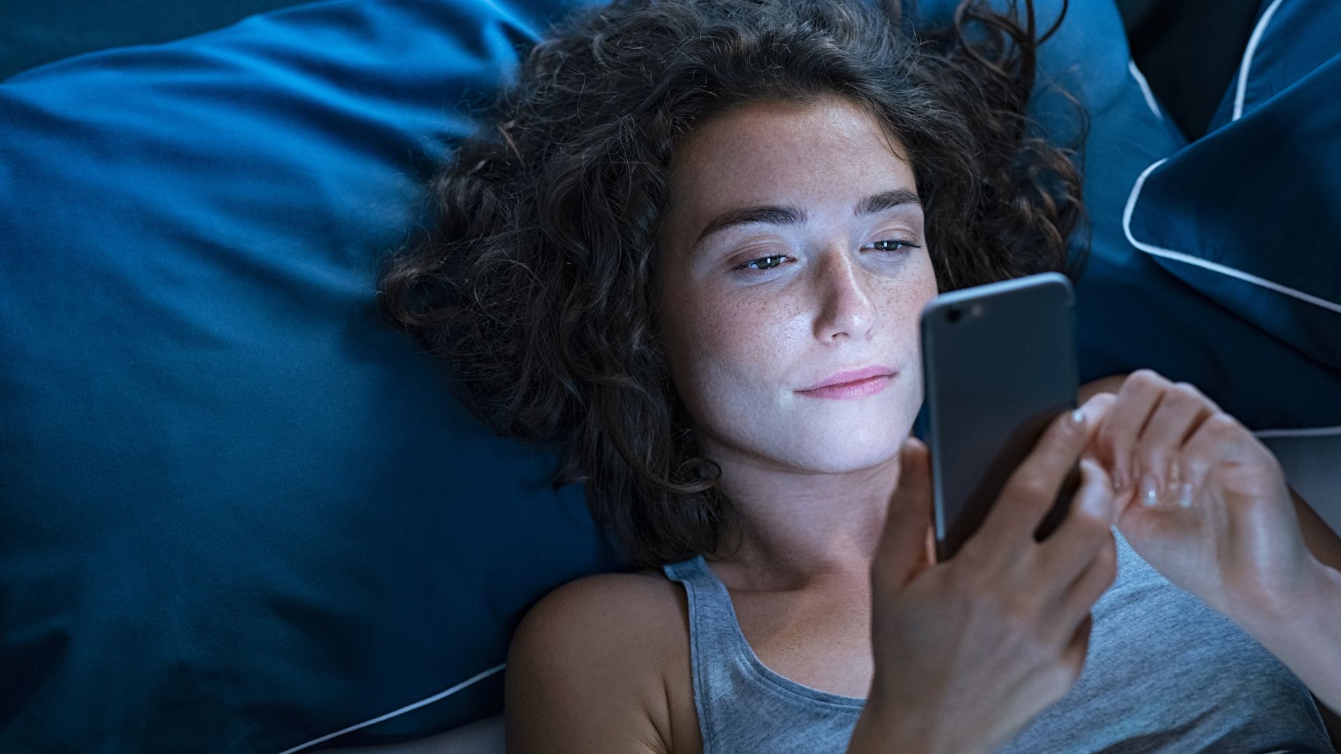 A woman lies in bed at night looking at her phone. The light from the screen illuminates her face.