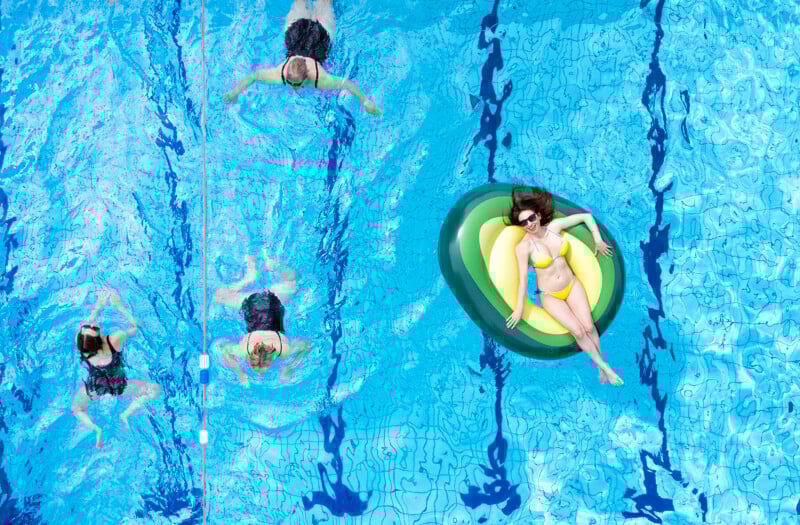 Four women are in a swimming pool; three swim in lanes while one in a yellow bikini relaxes on a green and yellow inflatable float, wearing sunglasses. The image is shot from above.