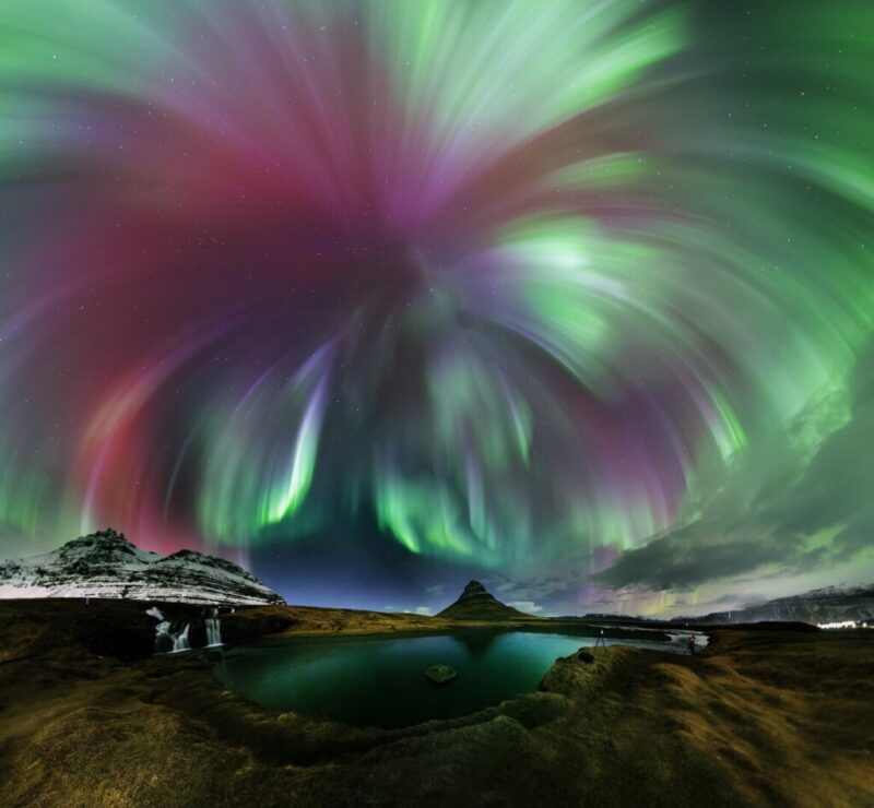 Northern lights photos: Green and red aurora seeming to emanate from one point and burst like a firework over a wide-angle landscape with mountains and a lake.