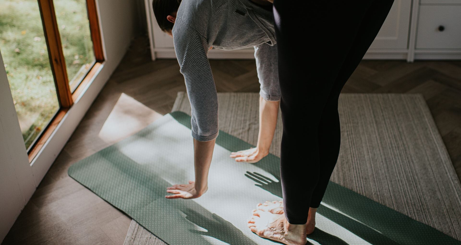 Woman doing standing Pilates workout in home studio, reaching down to touch yoga mat on the floor