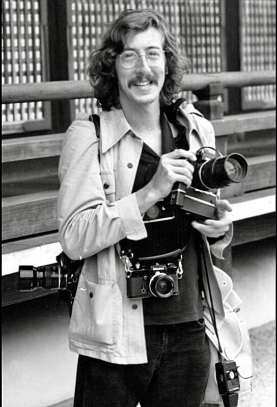 A smiling man with wavy hair and glasses holds a camera. He has two more cameras hanging around his neck and another on his shoulder. He wears a light jacket and stands outdoors near a wooden structure.