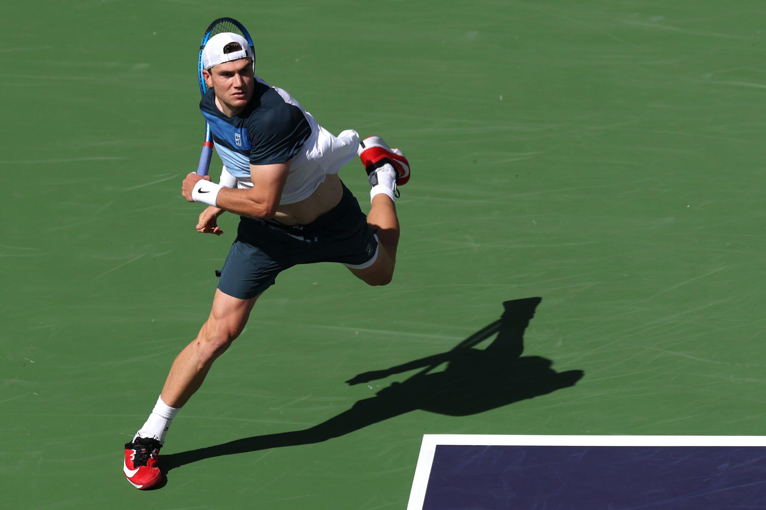 Jack Draper follows through on a forehand on a navy blue tennis court against a green backdrop.