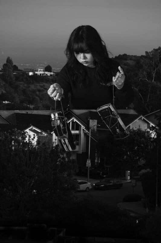 A black-and-white image shows a giant woman looming over suburban houses, manipulating two toy cars with strings like a puppeteer. The scene combines an outdoor neighborhood with a surreal, oversized figure.