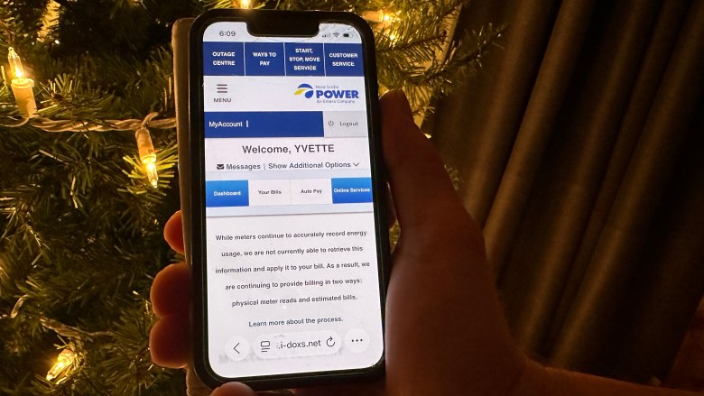 A hand holds a cellular phone in front of a Christmas tree with white lights. The phone is open to a Nova Scotia Power app and says "Welcome Yvette. While meters continue to accurately record energy usage, we are not currently able to retrieve the information and apply it to your bill. As a result, we are continuing to provide billing in two ways: physical meter reads and estimated bills."
