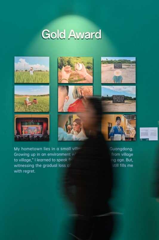 A blurred person walks past a green wall displaying the "Gold Award" title, a collection of nine photos, and a caption describing life in a small Guangdong village.