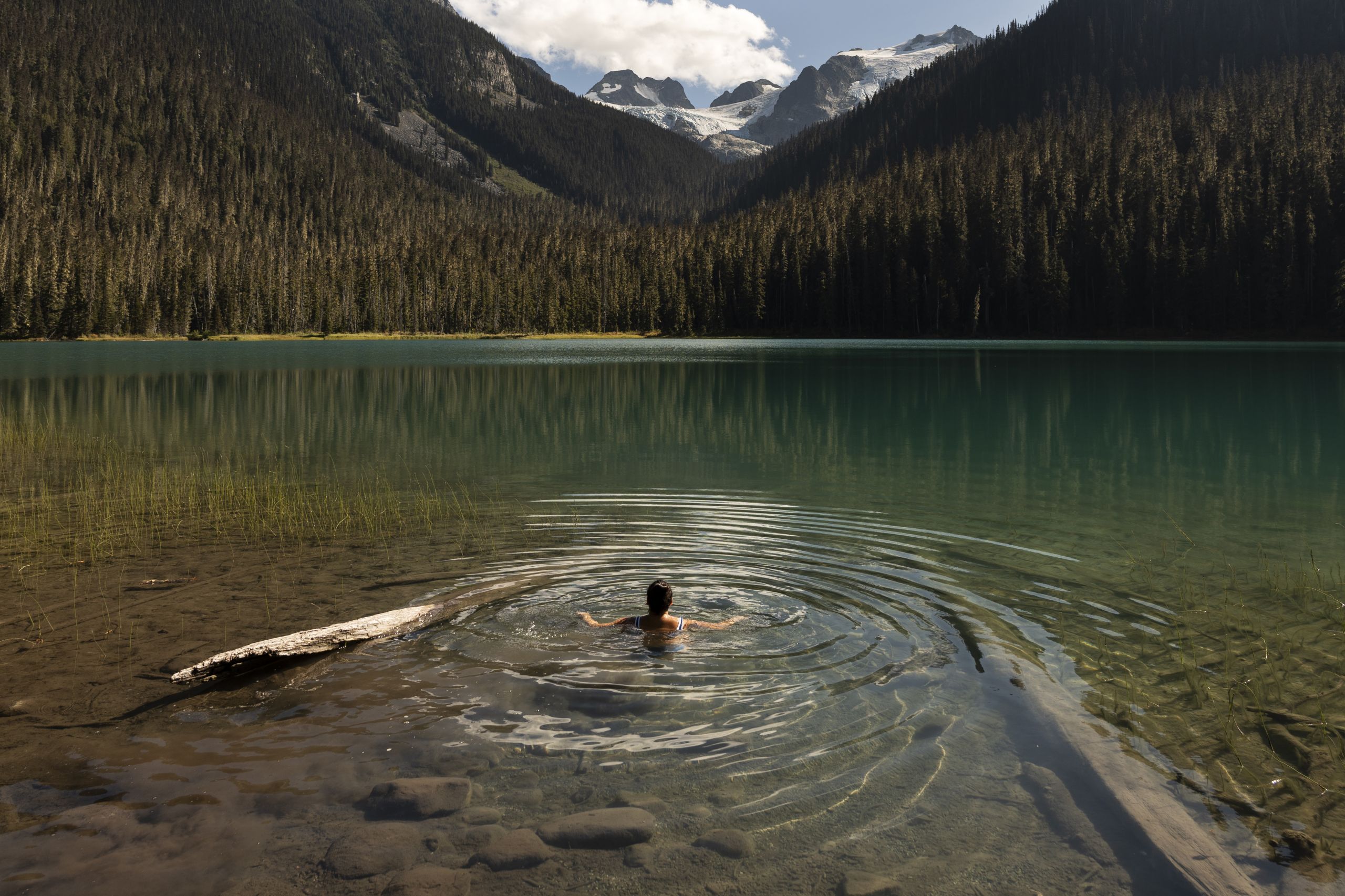 A person swims in a placid glacial lake with a scenic view of mountains, forest and sky on the other side
