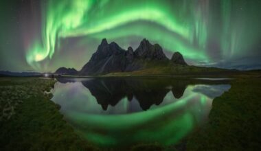 Swirling green aurora over sharp mountains with a reflection in a foreground lake.